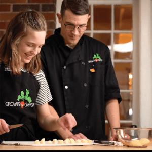 Andrew Fleet and his daughter Orla Fleet, both wearing Growing Chefs aprons, standing side by side while chopping vegetables in a kitchen.