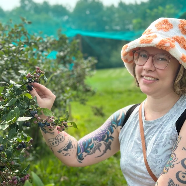A person with glasses and a white bucket hat with orange flowers is standing in a green outdoor setting, picking blueberries from a bush. They have tattoos on their arms and are wearing a light-colored tank top and a crossbody bag. The background shows more bushes and a netted structure.