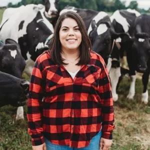 Janan Dean standing in front of a heard of dairy cows. Wearing a red buffalo plaid shirt Smiling with brown hair.