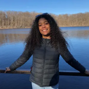 A smiling woman with curly hair, wearing a black jacket, standing in front of a lake with bare trees in the background.