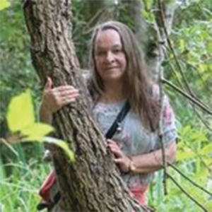 Maya Clarke, standing outdoors in a forest, leaning gently against a tree with a calm expression, surrounded by green foliage.