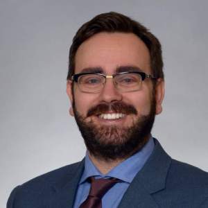 A smiling person with dark hair, a moustache and neatly trimmed beard. Wearing a dark blue blazer, lighter blue shirt, and a dark coloured tie. Grey background.