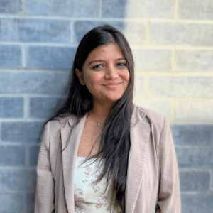 Smiling person with long dark hair. Standing in front of a brick wall. Wearing a pinkish-beige blazer and a white shirt.