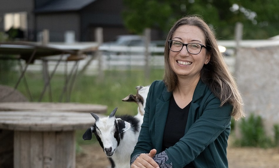 A smiling woman with long brown hair and glasses in a farm setting. She wears a green blazer, and a black shirt. Behind her, two goats are peaking out. The background is a fenced-in area with grass and trees.