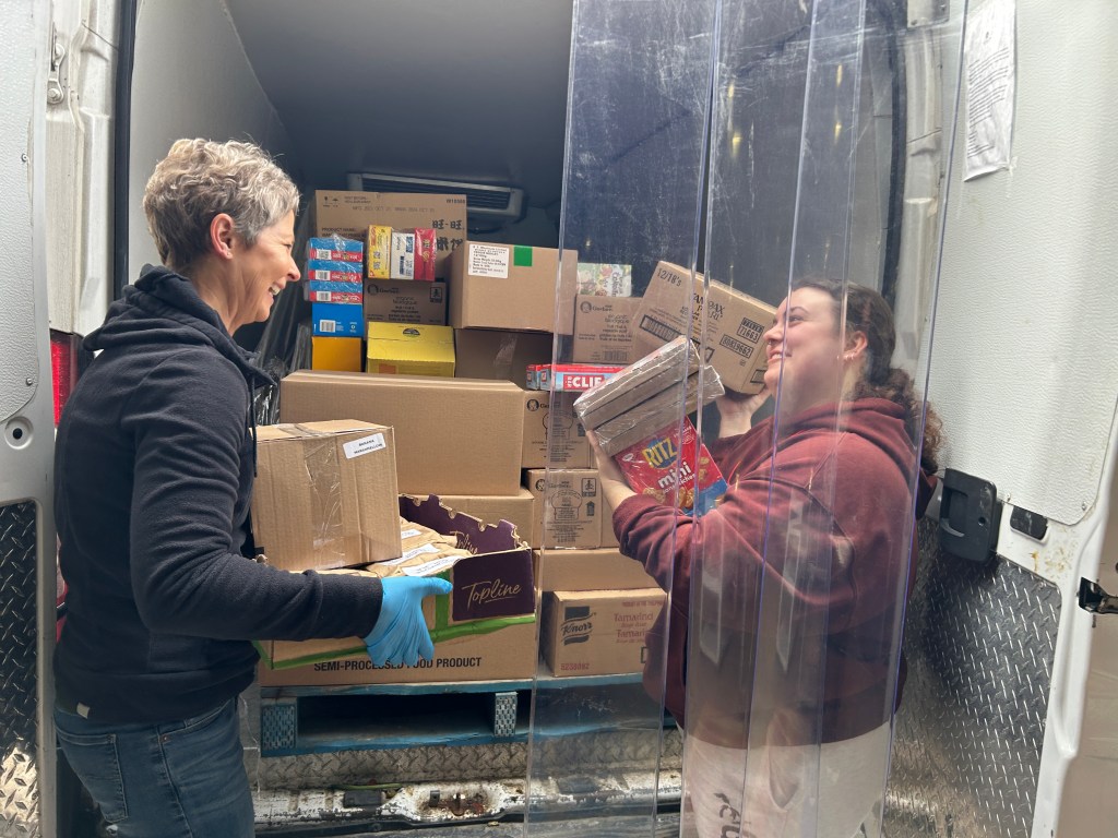 Two people are smiling while unloading boxes of food from the back of a truck. One person wearing gloves is carrying a box, while the other, in a maroon hoodie, is holding packages of crackers. The truck is filled with stacked food boxes.