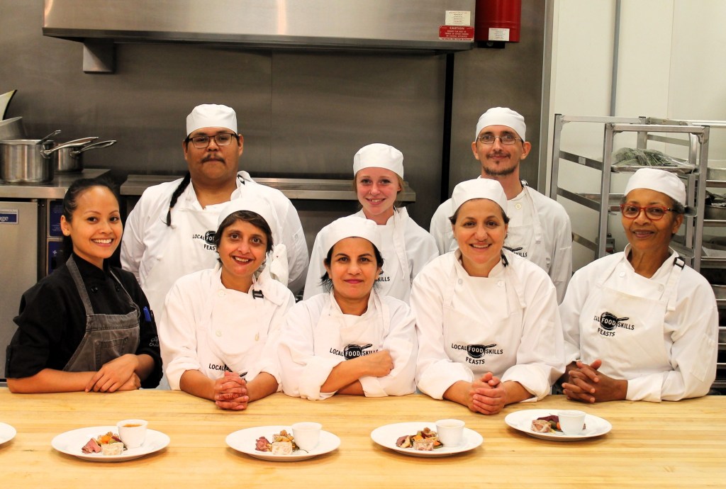 A group of eight chefs in a commercial kitchen.  Chandanny is on the left of the photo in a black shirt and dark apron.  The other learners are in white jackets and white hats.  There are is a wood counter in front of them with 4 plates.  
