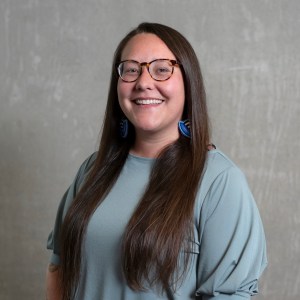 A smiling person with long dark hair, glasses. Standing in front of a grey background.