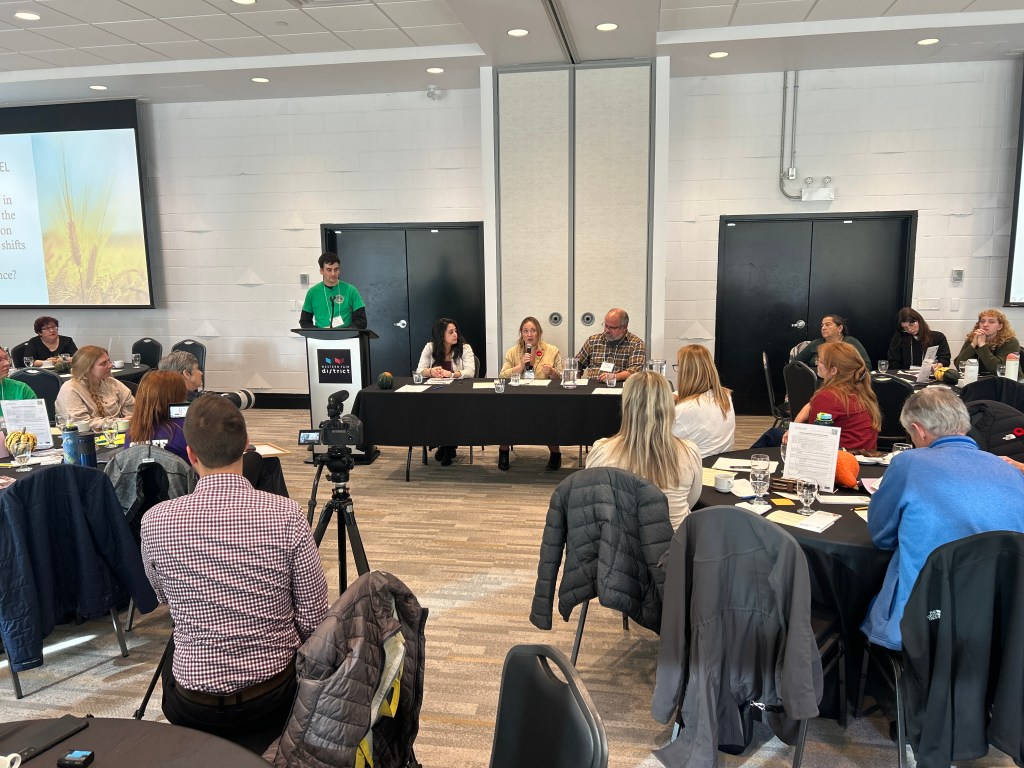 A panel discussion at featuring Michelle Stranges, Chantel Crockett, and Patrick Dunham seated at a long table with microphones and nameplates. A young man in a green shirt stands at the podium addressing the audience. Behind him, a large screen displays a presentation slide with an image of wheat. Attendees are seated at round tables, listening and taking notes, while a camera records the event.