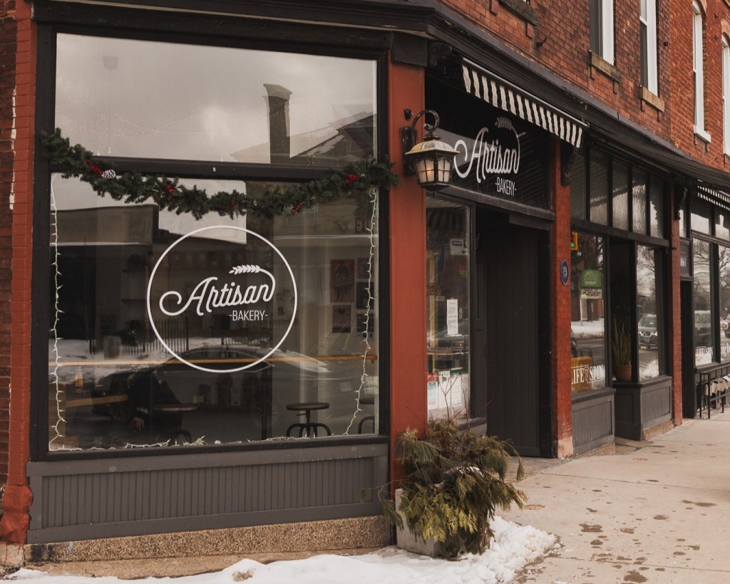 Exterior of a cozy brick bakery on a snowy street, featuring large glass windows with a festive garland and string lights. The window displays a white logo reading “Artisan Bakery” with a stylized wheat graphic. A black-and-white striped awning and vintage lantern frame the entrance, with evergreen foliage in a sidewalk planter.