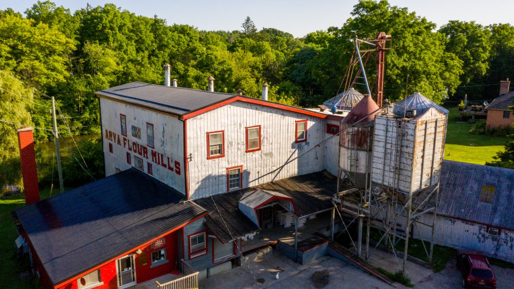 Aerial view of historic Arva flour mill with white and red wooden buildings, attached silos, and metal conveyors, surrounded by green trees and lawns on a sunny day.