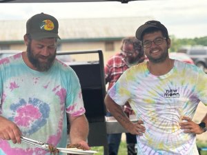 Two people wearing tie-dye shirts stand at an outdoor grill, with one using tongs to plate food. A building and open farm area are visible in the background.