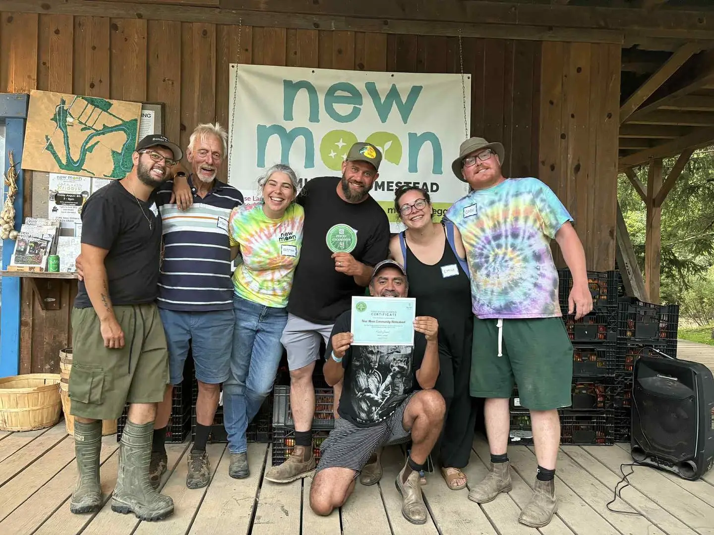 Group of seven standing on a wooden stage beneath a “new moon” homestead banner, one person kneeling and holding a certificate.