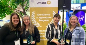 Four individuals stand together holding glass awards in front of a large banner that reads “Ontario – Excellence in Agriculture Program.” The group is positioned inside an event space with greenery and display booths visible in the background
