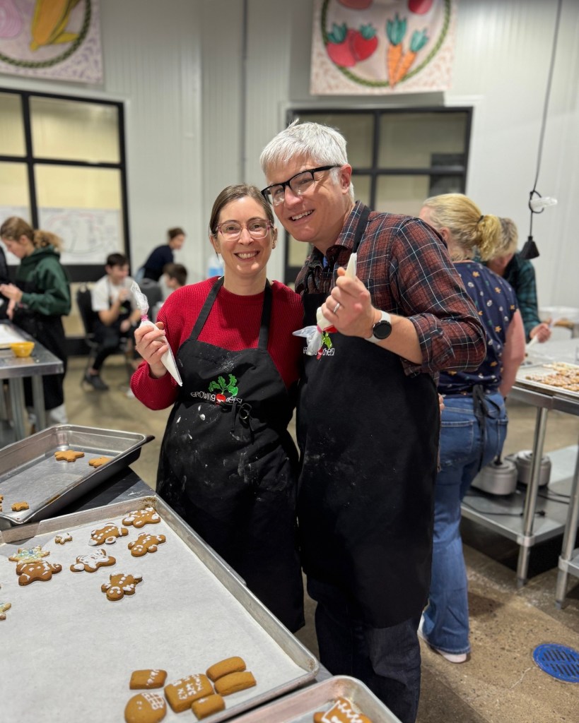 Two individuals wearing aprons stand together in a baking workspace, each holding a piping bag. They are surrounded by trays of undecorated and partially decorated gingerbread cookies on stainless steel tables. Other people in the background are also working on cookie decorating. Colorful artwork hangs on the wall above large windows.