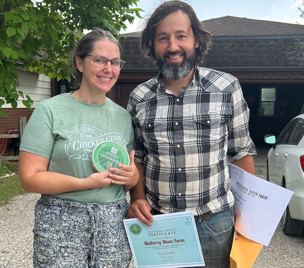 Two individuals stand outside a farm building holding certificates and an award plaque. One certificate reads “Mulberry Moon Farm.” They are positioned in front of a driveway with a tree and parked vehicles visible in the background.