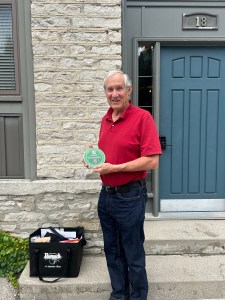 Crispin Colvin stands outside a stone building beside a blue door marked “18,” holding the 2025 Food System Champion green circular plaque, with a black tote bag at his feet.