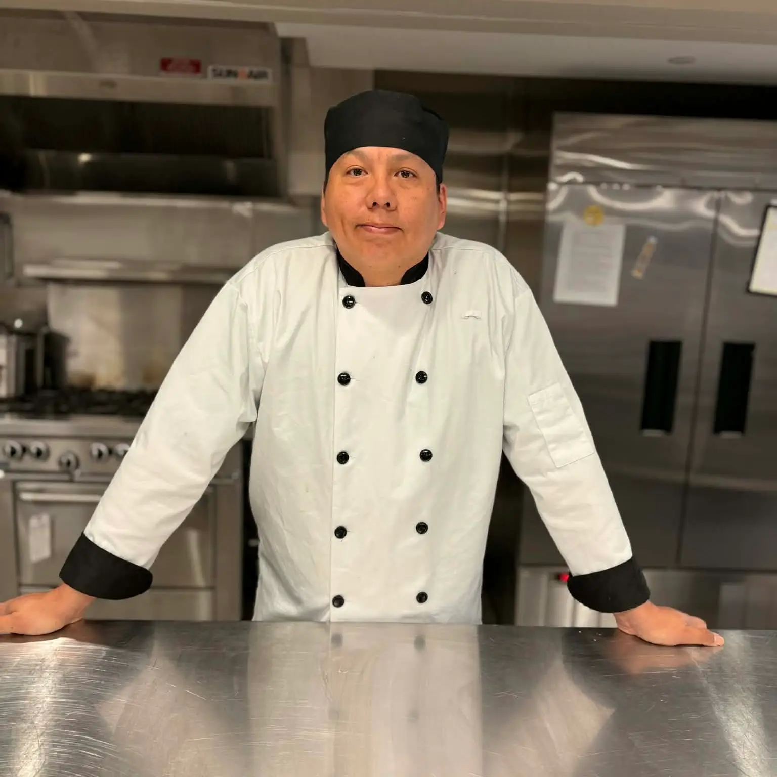 Shawn in an industrial kitchen wearing a white chefs jacket with black buttons and cuffs. Wearing a black low profile chefs hat.
