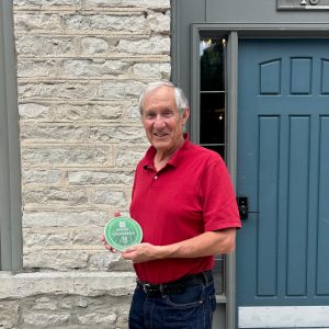 A man in a red shirt stands holding a small green plaque. There is a blue door in the background