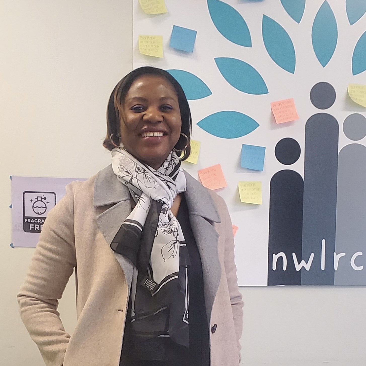 Rachael Bem Sai standing indoors at the Northwest London Resource Centre, with a community message board featuring colourful notes and a tree graphic behind her.
