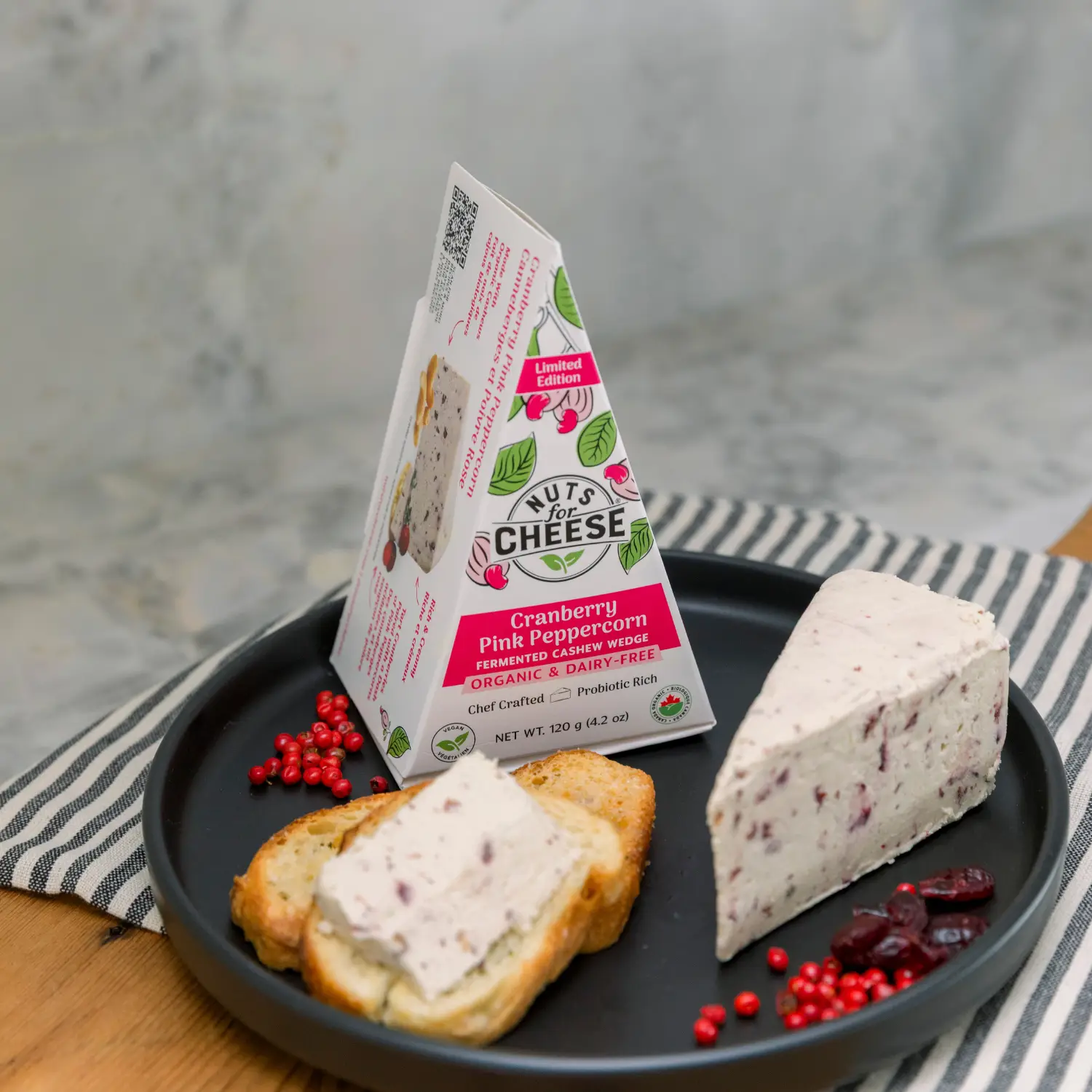 Cranberry pink peppercorn fermented cashew cheese wedge with packaging, served on toast on a black plate.
