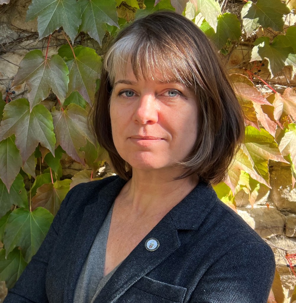 Head-and-shoulders portrait of a Justine Dainard wearing a dark jacket, standing outdoors in front of a leafy ivy-covered wall.