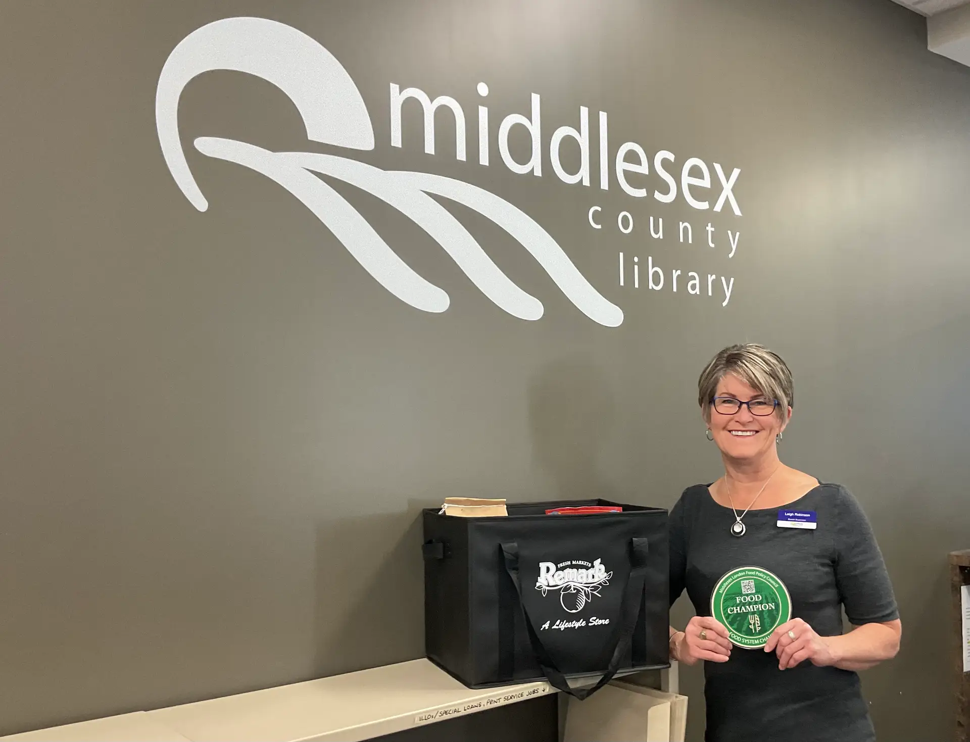 A person stands inside a Middlesex County Library branch beside a reusable grocery tote and holds a circular "Food Champion" sign. The wall behind them displays the Middlesex County Library logo. The image highlights a partnership between the library and local food system initiatives.