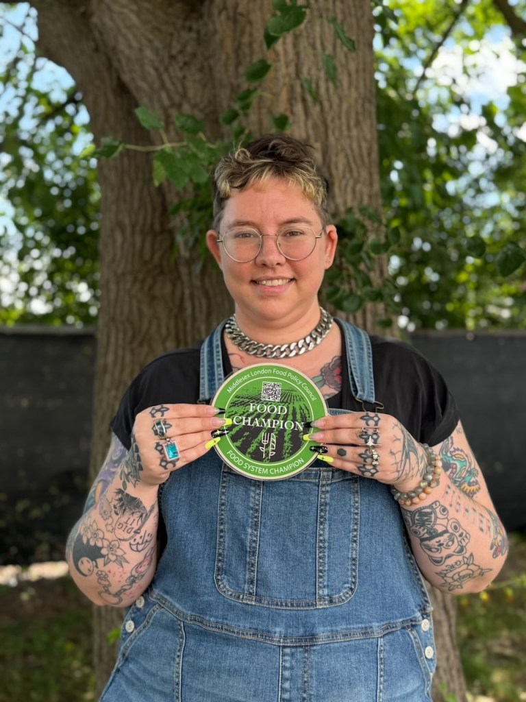River Rush, smiling and wearing denim overalls. Holding MLFPC Food System Champion plaque. Standing in front of a tree with leaves.