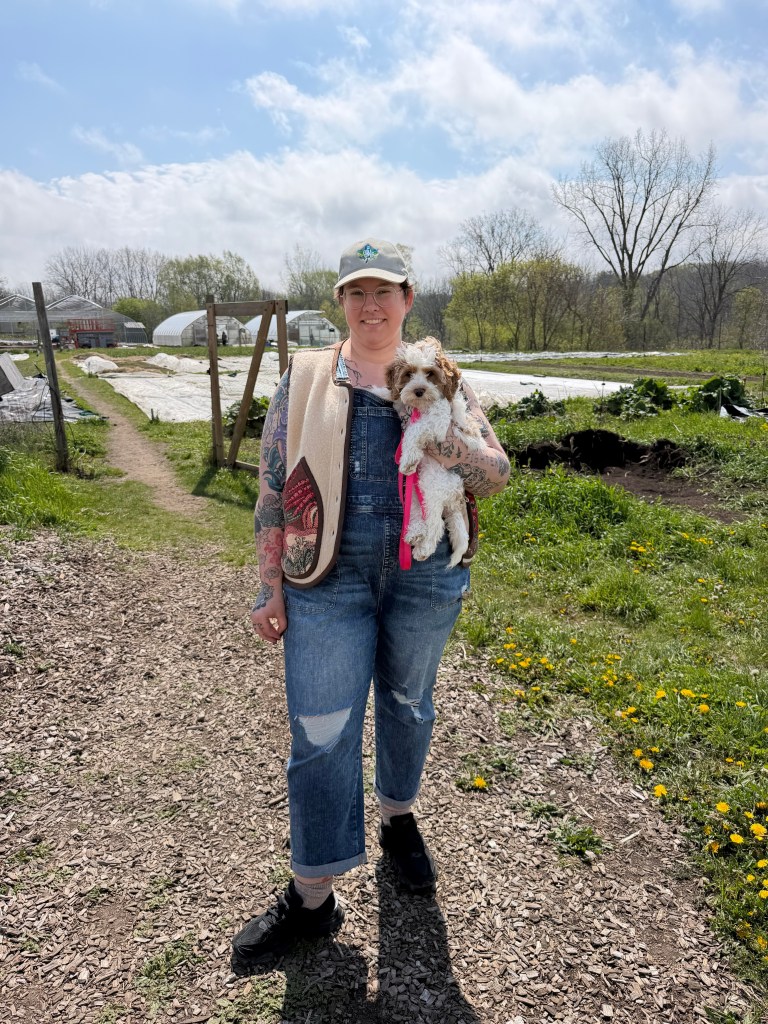 River Rush in an agriculture setting. Wearing a hat and colourful jack and overalls. Holding a small brown and white dog.