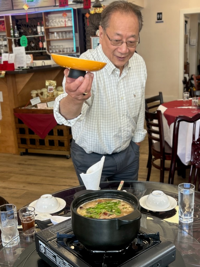 Dr. Ed Lui standing over table, holding a pot lid. on th table is a pot of rich delicious soup.