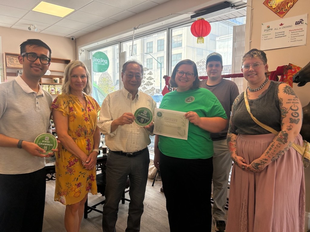 A group of size people standing in a line. In the middle of the photo, Dr. Ed Lui holds a MLFPC Food Champion plaque and MLFPC Food System Facilitator Kathy MacKay presents Dr. Lui with a certificate.