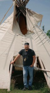 Kyle Albert stands at the entrance of a traditional white tipi, wearing a black shirt and jeans, at Nimkee Nupigawagan Healing Centre.