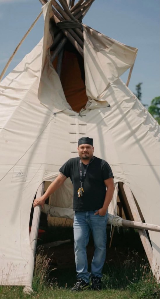 Kyle Albert stands at the entrance of a traditional white tipi, wearing a black shirt and jeans, at Nimkee Nupigawagan Healing Centre.