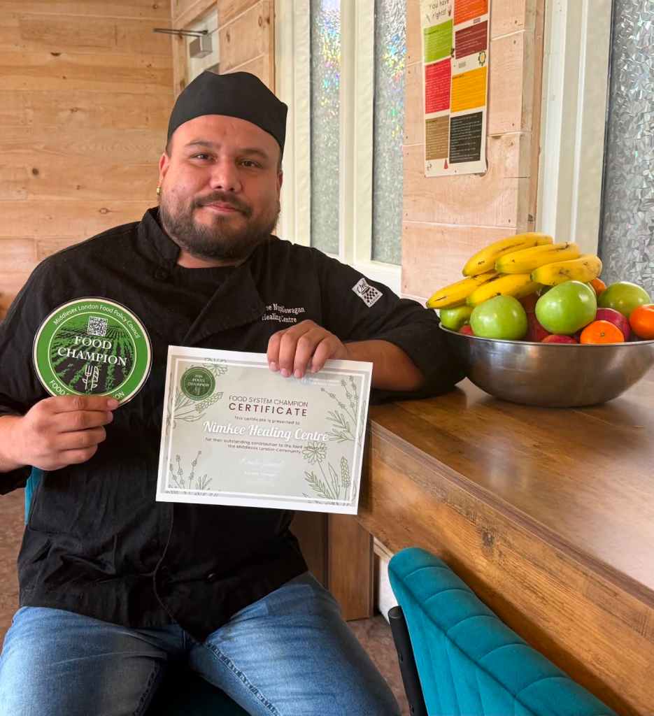 Kyle Albert sits beside a bowl of fresh fruit, holding a Middlesex-London Food Policy Council Food Champion sticker and a Food System Champion certificate recognizing Nimkee Nupigawagan Healing Centre.